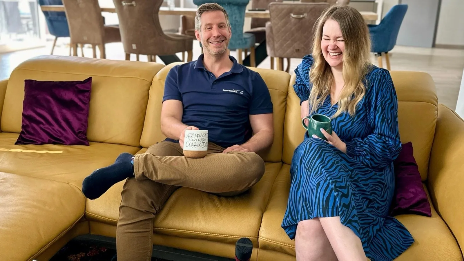 Sascha Mewes-Herzog (links) und seine Frau Sarah (rechts) sitzen auf einem gelben Sofa und lachen.