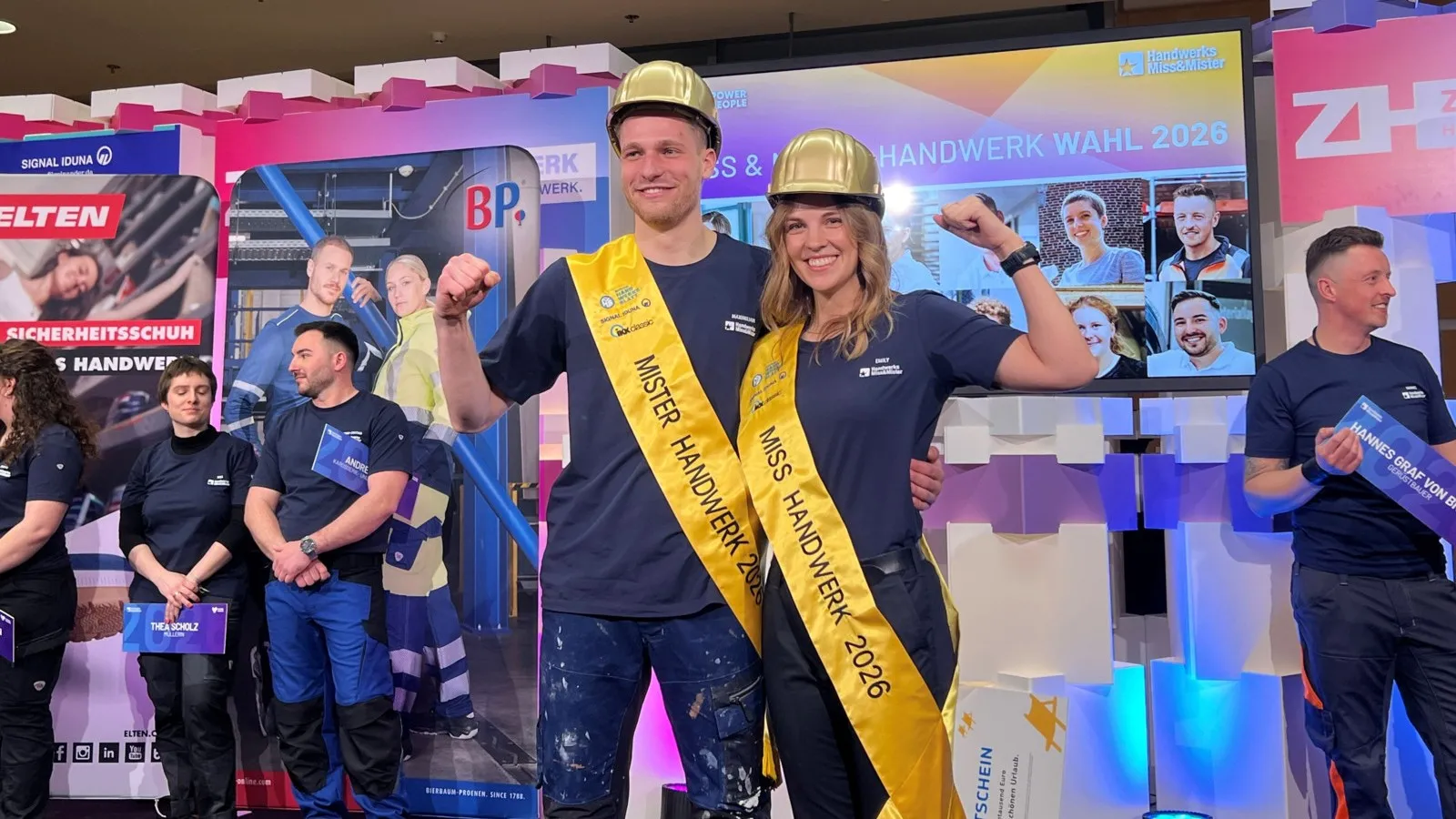 Emily Maichle und Maximilian Buerger stehen nebeneinander. Sie tragen eine gelbe Schärpe und einen goldenen Helm und freuen sich über ihre Auszeichnung.