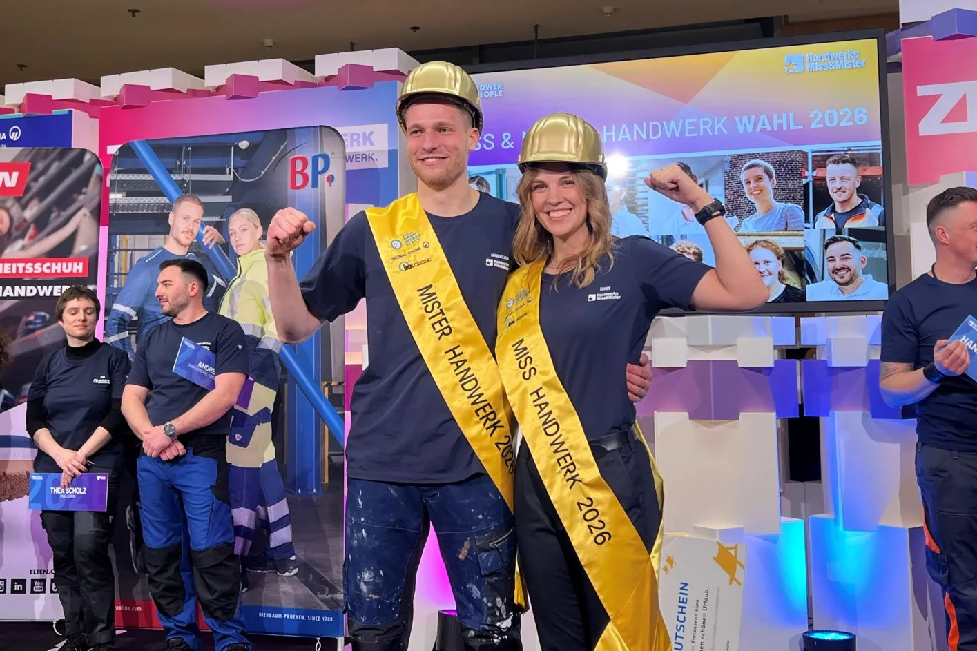 Emily Maichle und Maximilian Buerger stehen nebeneinander. Sie tragen eine gelbe Schärpe und einen goldenen Helm und freuen sich über ihre Auszeichnung.