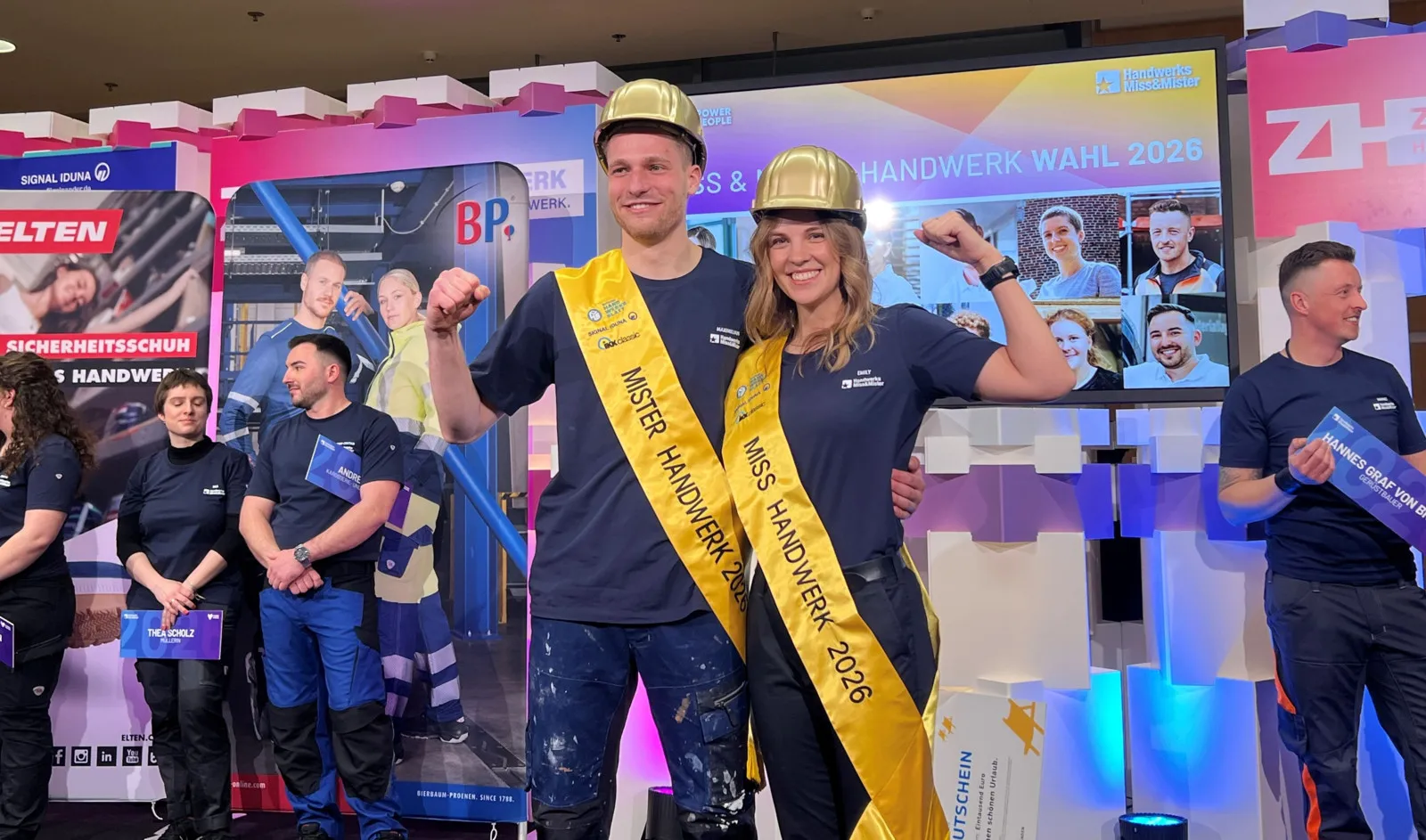 Emily Maichle und Maximilian Buerger stehen nebeneinander. Sie tragen eine gelbe Schärpe und einen goldenen Helm und freuen sich über ihre Auszeichnung.