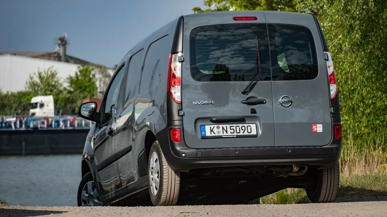 Der grau-schwarze Nissan NV250 von hinten, an einem See stehend.