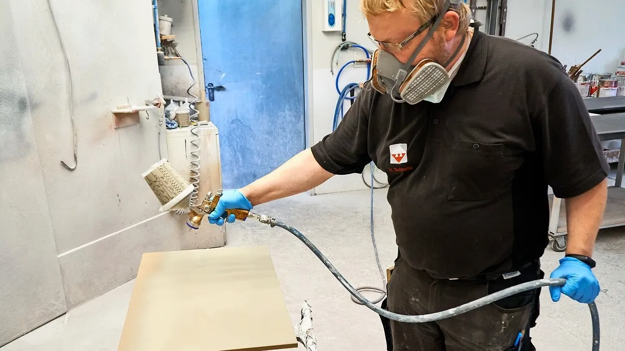 Tischlergeselle Hanno Schnelle mit Mundschutz benutzt die Satajet 1000 B Lignum 2. Er besprüht in einer Werkstatt eine Holzplatte.