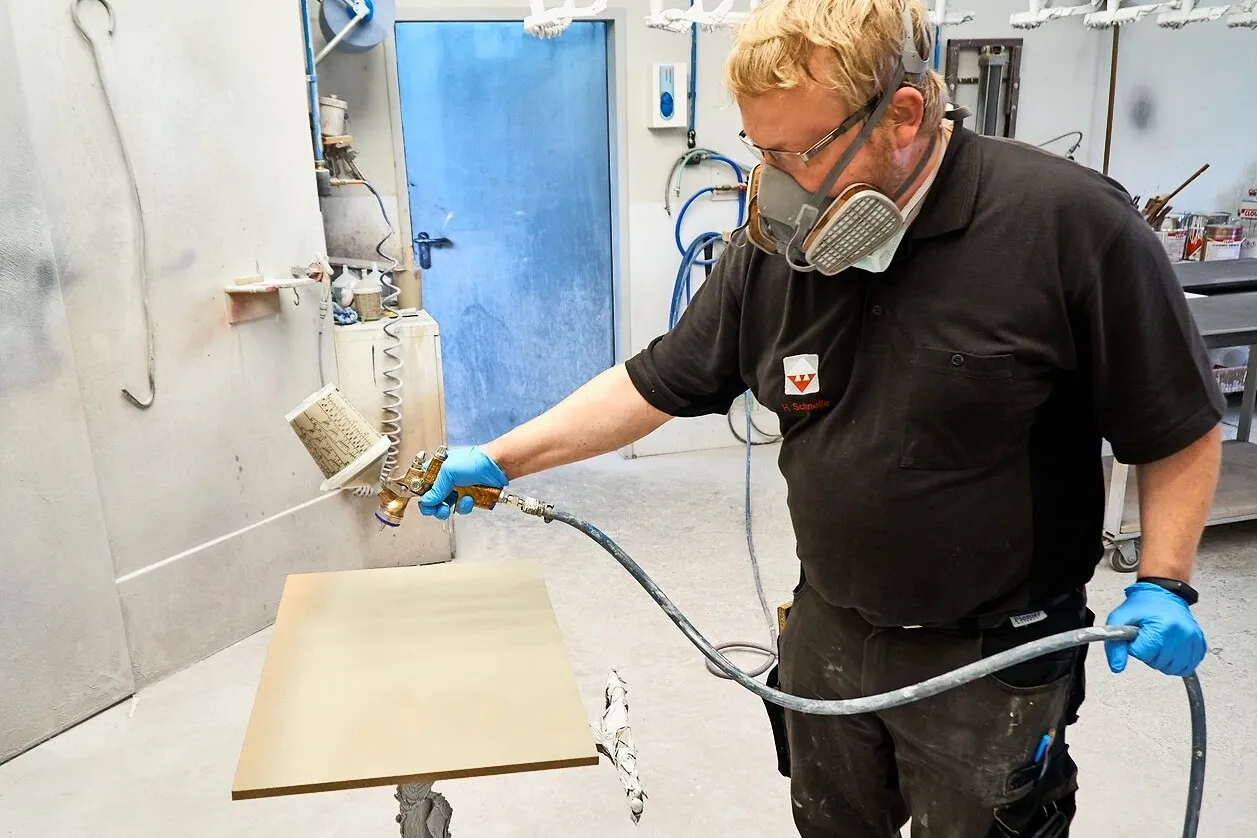 Tischlergeselle Hanno Schnelle mit Mundschutz benutzt die Satajet 1000 B Lignum 2. Er besprüht in einer Werkstatt eine Holzplatte.