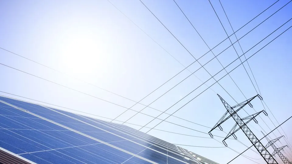 Eine Solaranlage im Vordergrund und ein Strommast im Hintergrund vor blauem Himmel.