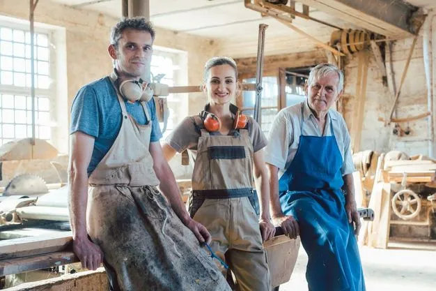 Drei Handwerker in Latzhosen stehen nebeneinander an einen Tisch gelehnt in einer Werkstatt und schauen in die Kamera.