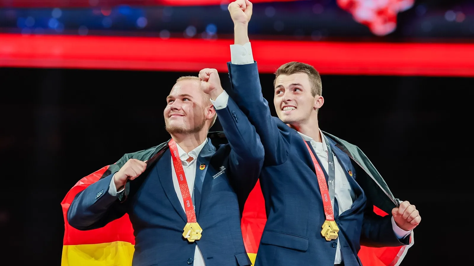 Tim Arnold und Jakob Distler jubeln mit den Fäusten in der Luft. Um ihre Schultern hängt die Deutschlandfahne. Um den Hals tragen sie die Goldmedaille.