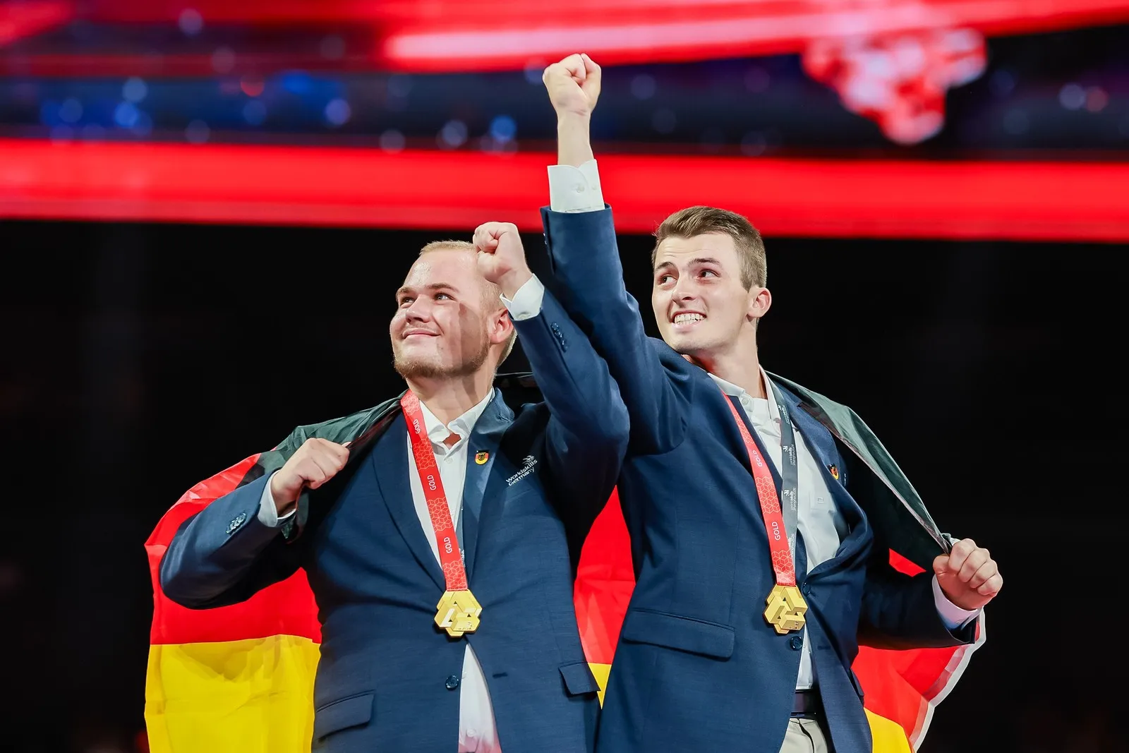 Tim Arnold und Jakob Distler jubeln mit den Fäusten in der Luft. Um ihre Schultern hängt die Deutschlandfahne. Um den Hals tragen sie die Goldmedaille.