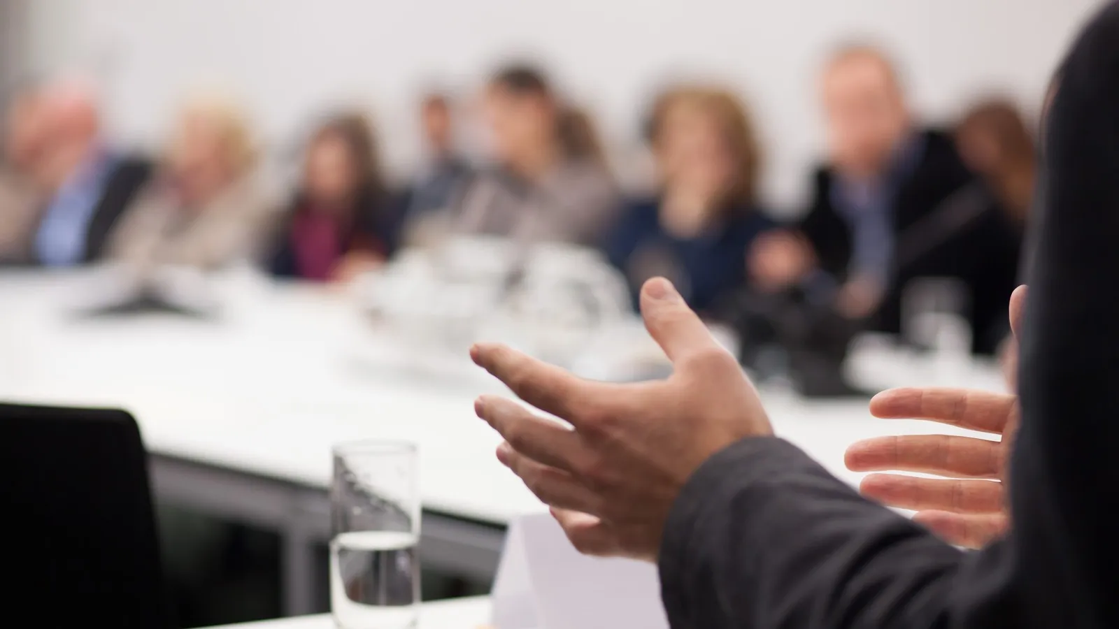 Mehrere Menschen sitzen in einem Konferenzsraum. Sie sind jedoch nur unscharf zu erkennen. Im Fokus befindet sich eine gestikulierende Hand.