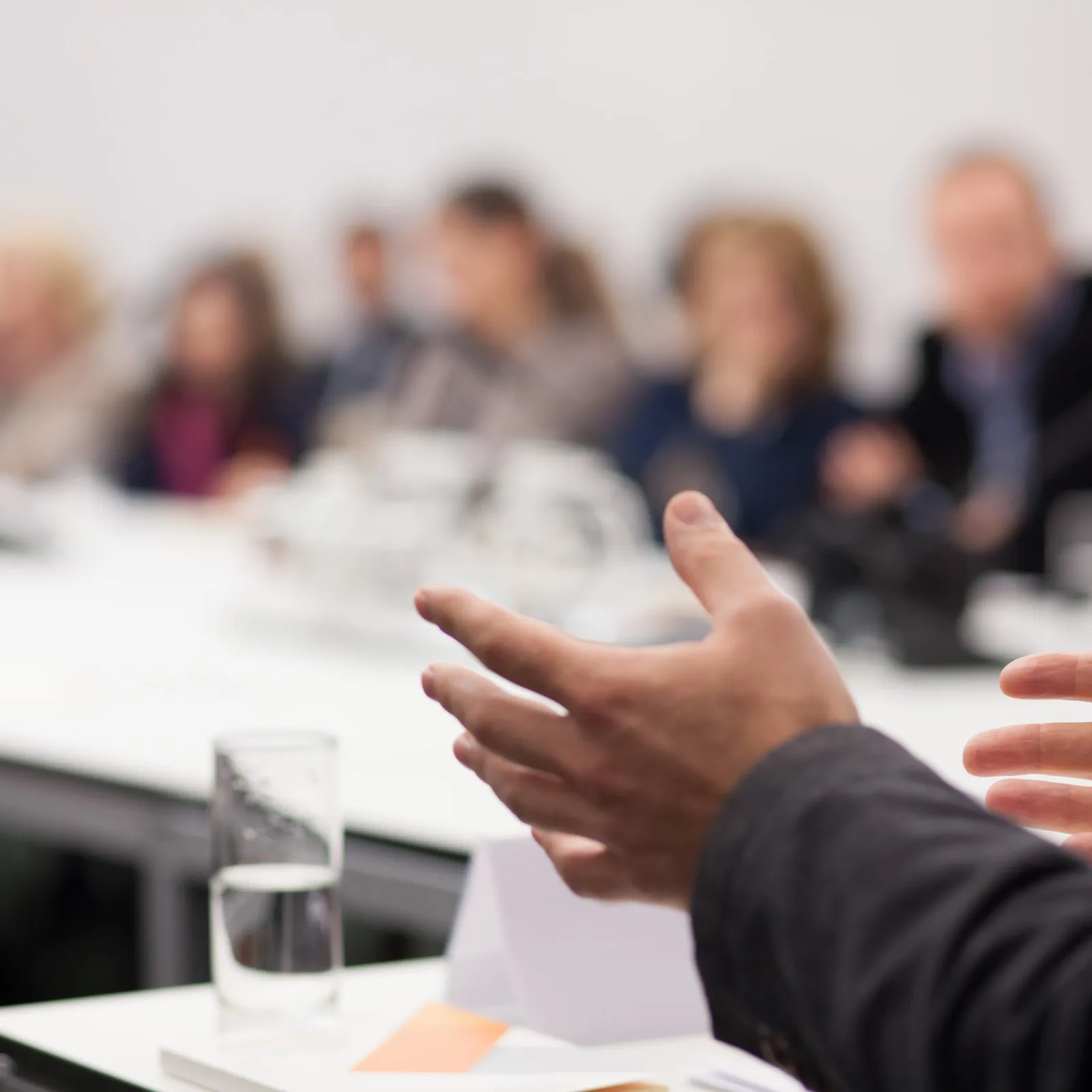 Mehrere Menschen sitzen in einem Konferenzsraum. Sie sind jedoch nur unscharf zu erkennen. Im Fokus befindet sich eine gestikulierende Hand.