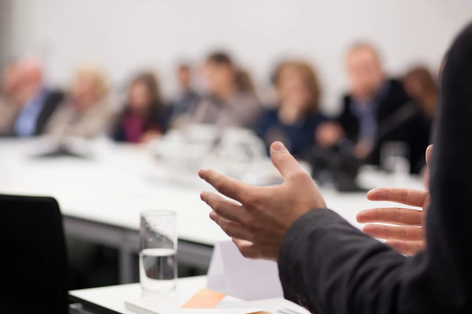 Mehrere Menschen sitzen in einem Konferenzsraum. Sie sind jedoch nur unscharf zu erkennen. Im Fokus befindet sich eine gestikulierende Hand.