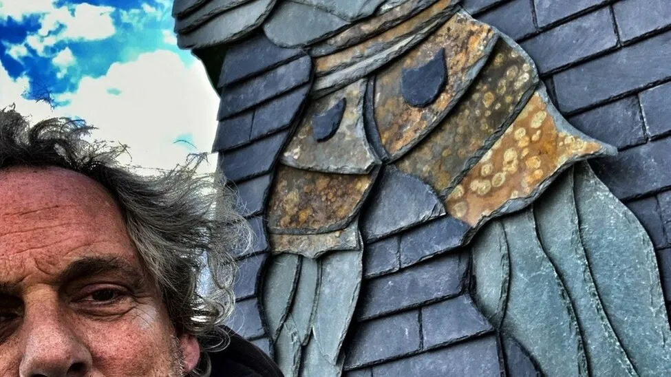 Michael Wenzel steht vor einem Dach, an dem er eins seiner Kunstwerke eingearbeitet hat.
