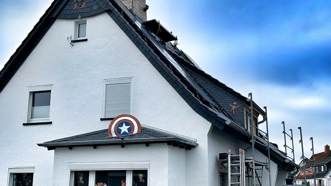 Ein weißes Haus aus der Frontansicht. Das Dach des Hauses hat vereinzelt Comic-Symbole eingearbeitet, wie z.B. vorne über dem Fenster das Schild von Captain America.