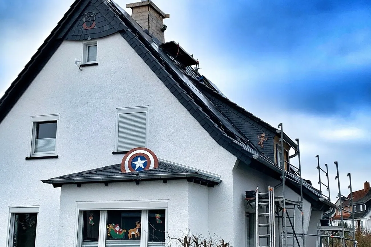 Ein weißes Haus aus der Frontansicht. Das Dach des Hauses hat vereinzelt Comic-Symbole eingearbeitet, wie z.B. vorne über dem Fenster das Schild von Captain America.