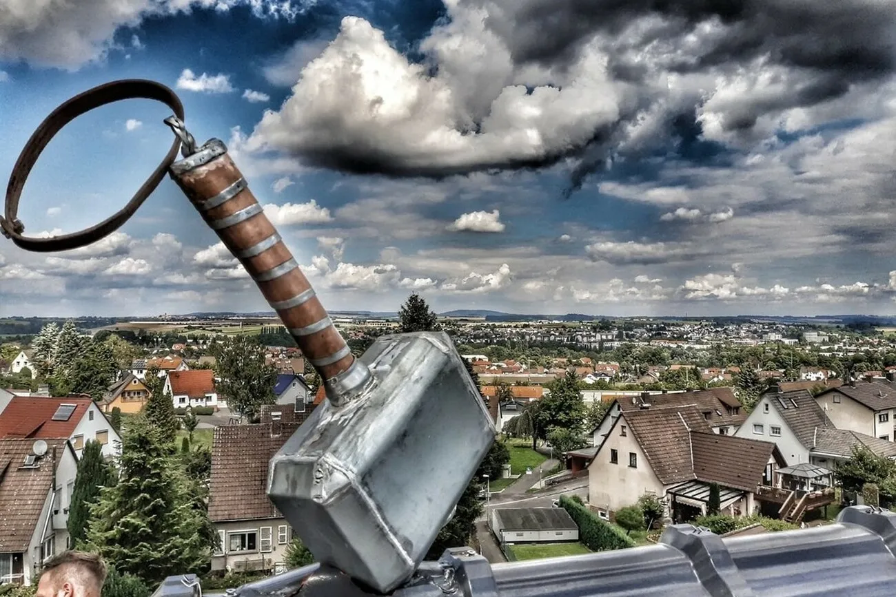 Nahaufnahme eines Daches, Im Fokus steht das Kunstwerk "Thors Hammer" mit vielen Häusern und Bäumen im Hintergrund.