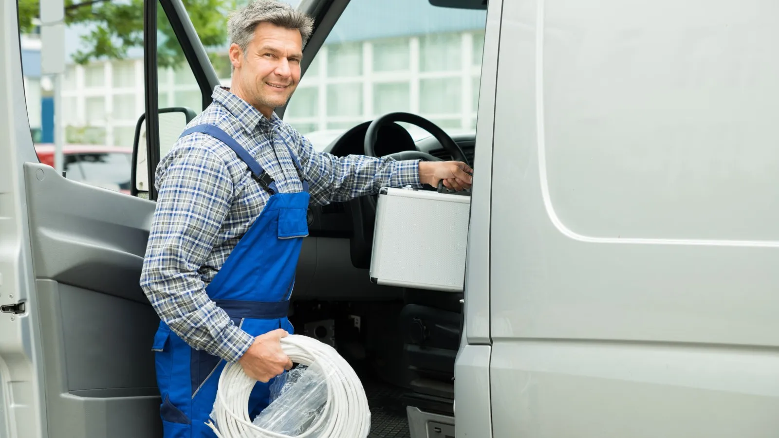 Ein Handwerker mit einer blauen Latzhose hält einen weißen Schutzhelm in der Hand und steht an einem weißen Transporter mit geöffneter Tür.