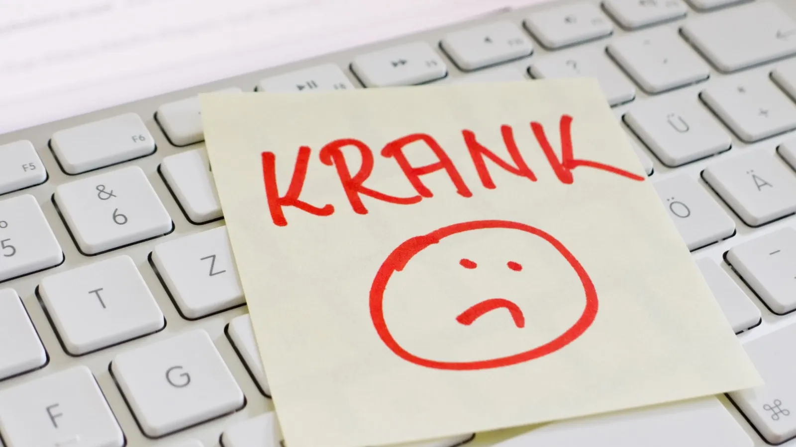 Ein Notizzettel mit der Aufschrift "KRANK" in rot und darunter ein trauriger Smiley. Der Zettel liegt auf einer weißen Computertastatur.