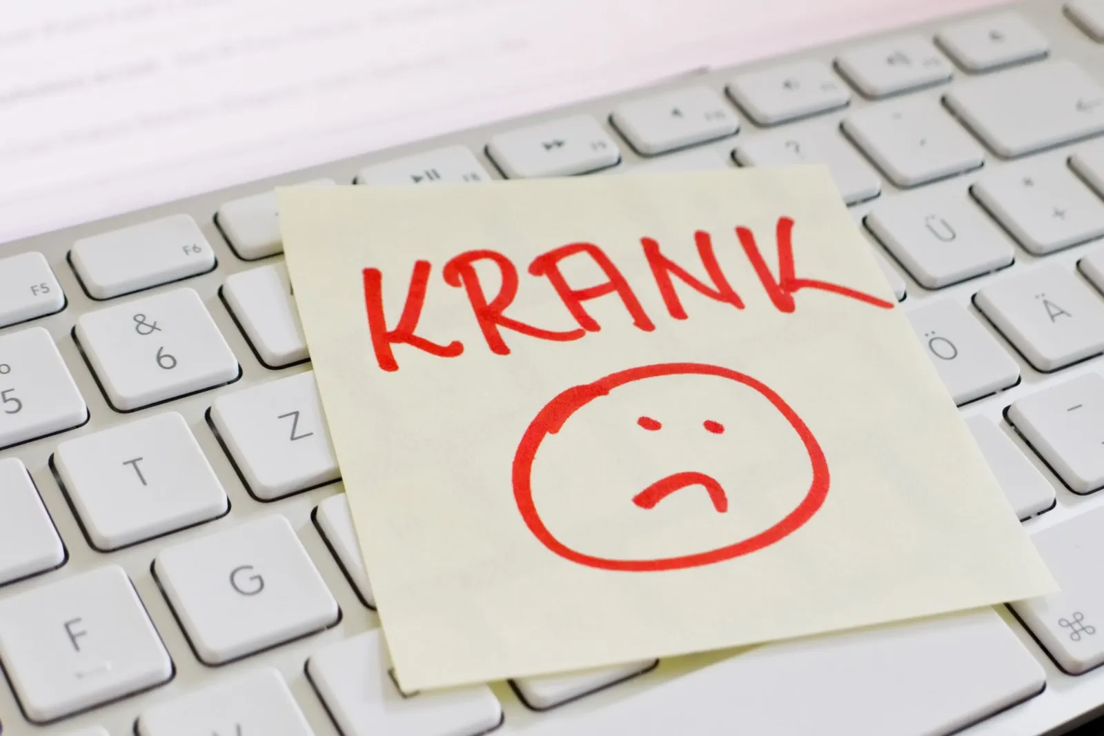 Ein Notizzettel mit der Aufschrift "KRANK" in rot und darunter ein trauriger Smiley. Der Zettel liegt auf einer weißen Computertastatur.