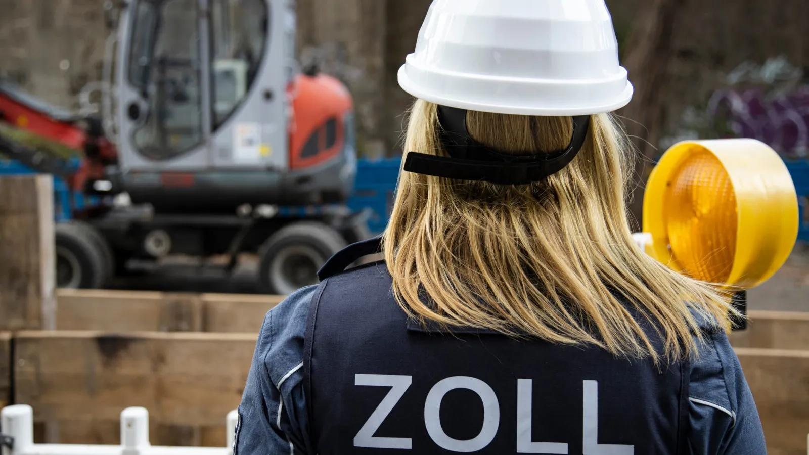 Eine Frau mit einem weißen Schutzhelm und einer Weste mit der Aufschrift "Zoll" ist von hinten zu sehen. Im Hintergrund ist eine Baustelle.