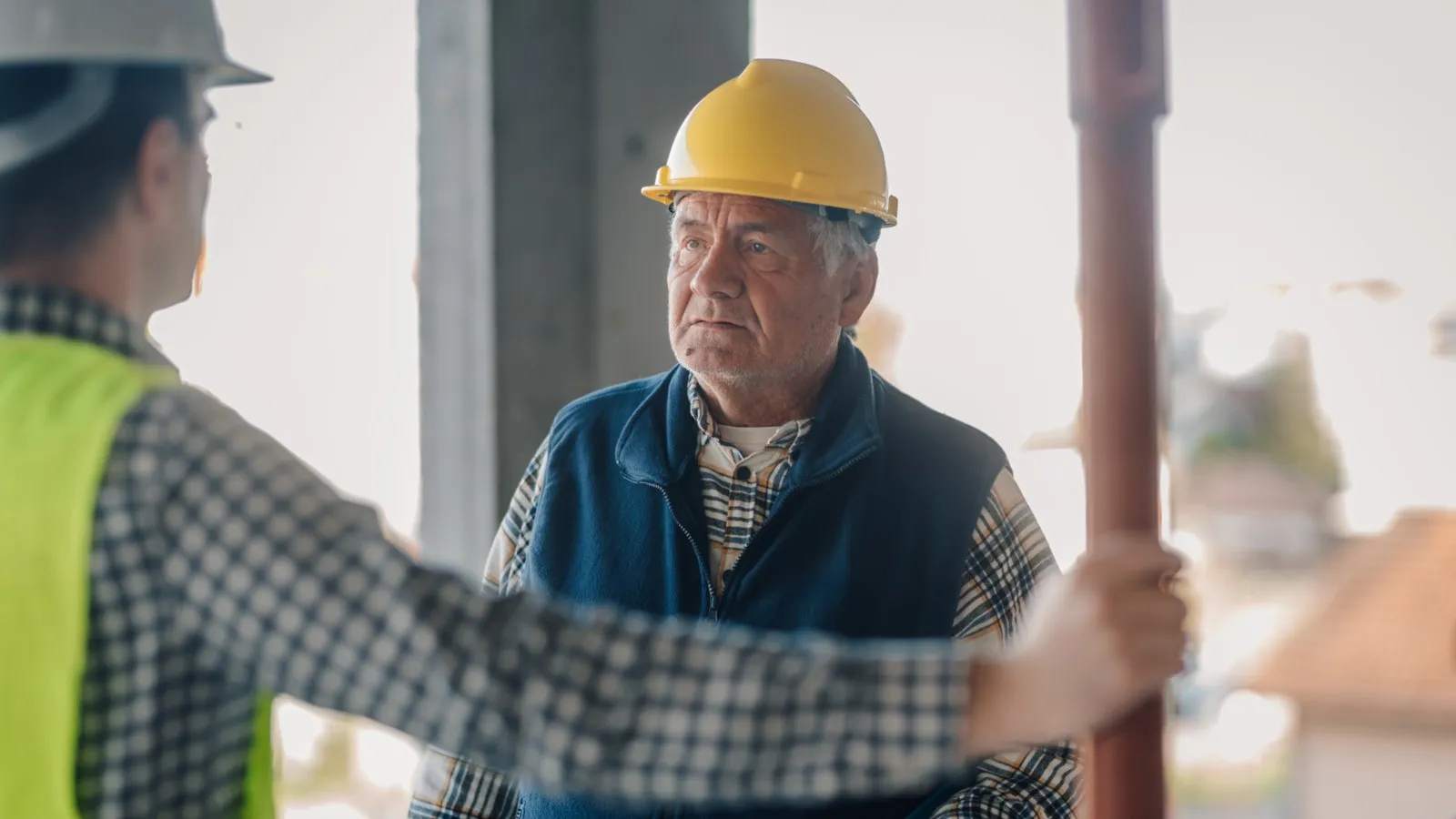 Ein älterer Herr mit gelbem Schutzhelm steht seinem Arbeitgeber gegenüber.