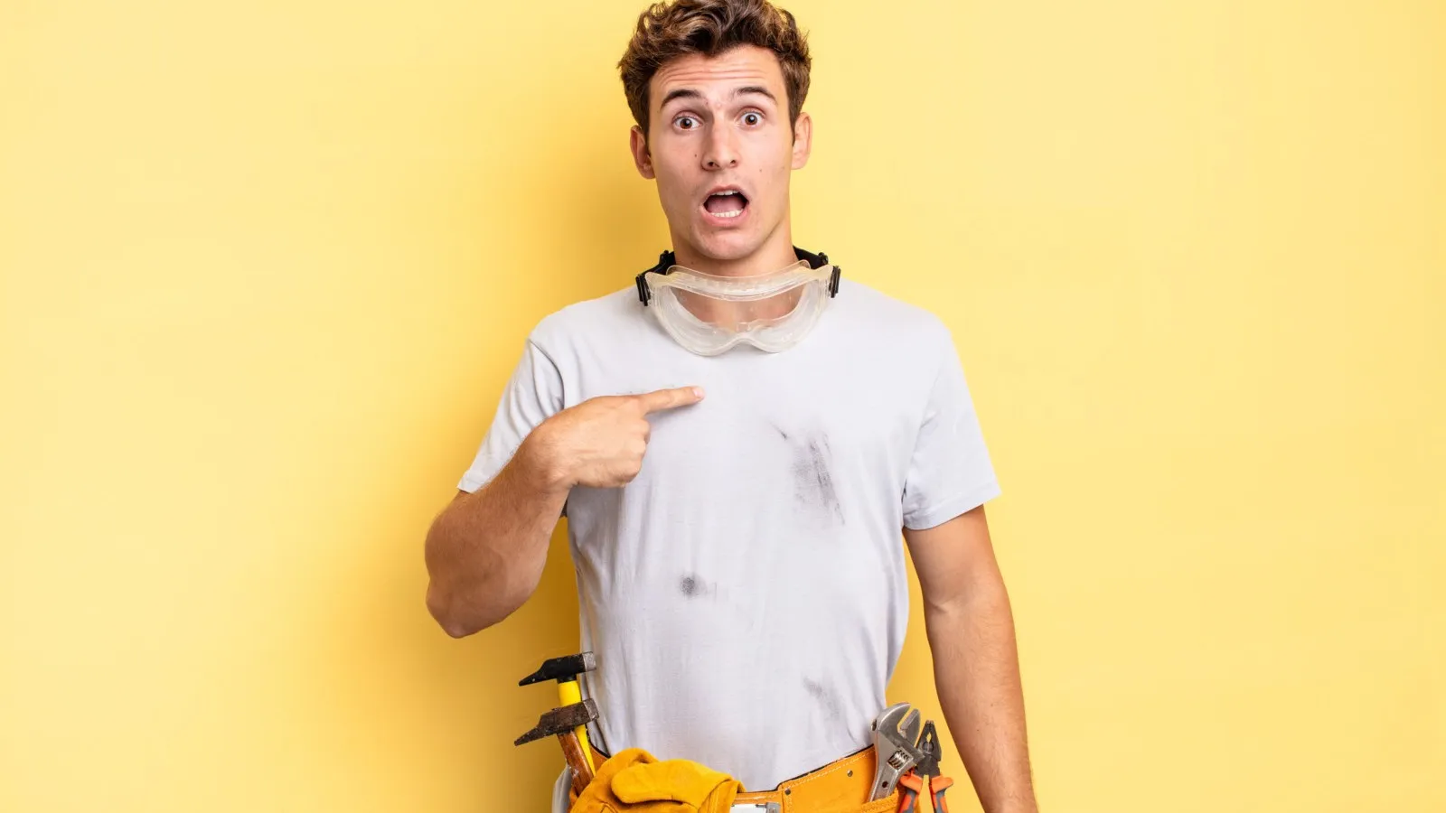 Ein überraschter Handwerker zeigt mit dem Finger auf sich. Er steht vor einem hellgelben Hintergrund.