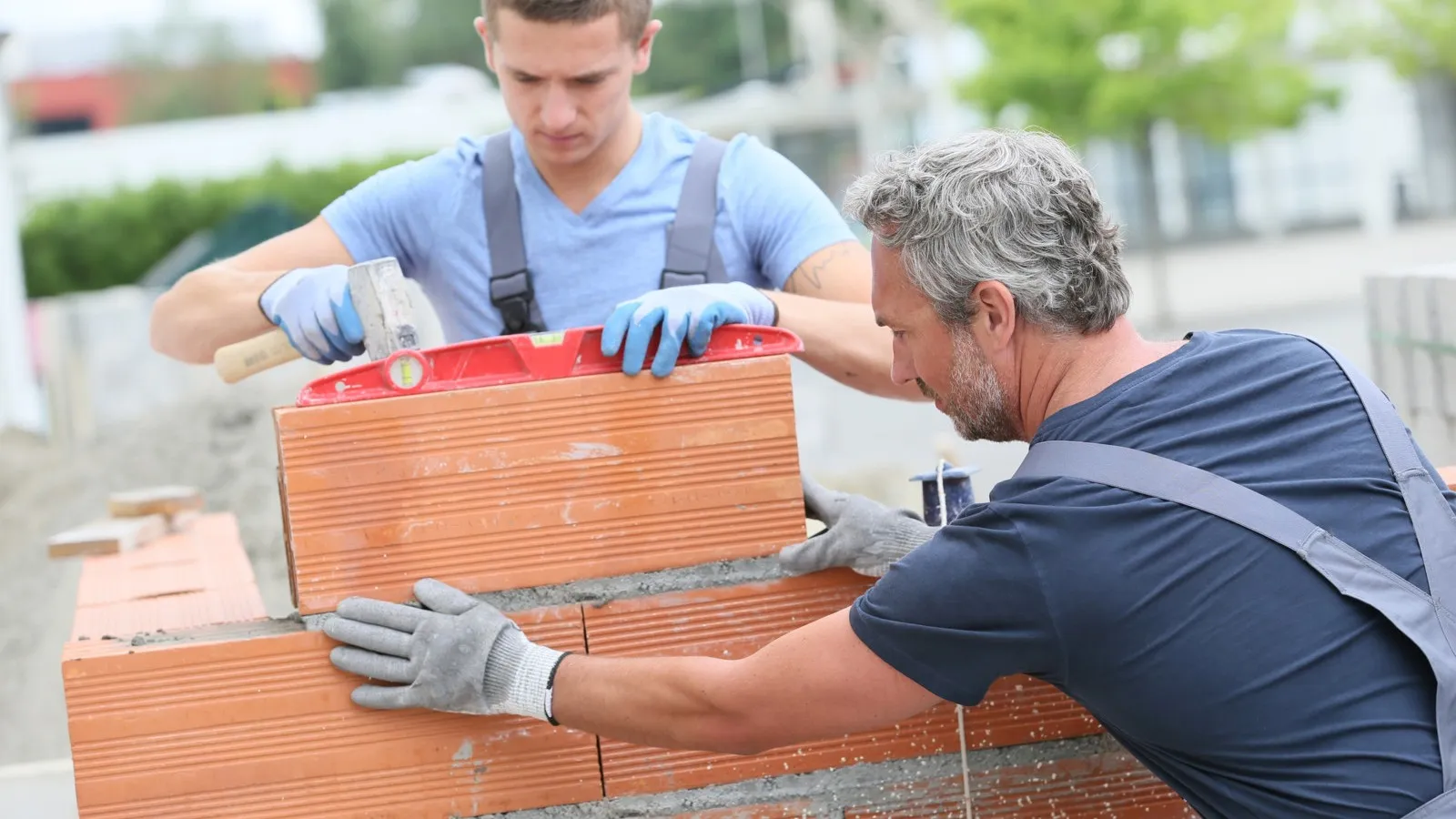 Zwei Handwerker, ein älterer im dunkelblauen T-Shirt und ein jüngerer in einem hellblauen T-Shirt arbeiten zusammen an einer Mauer.
