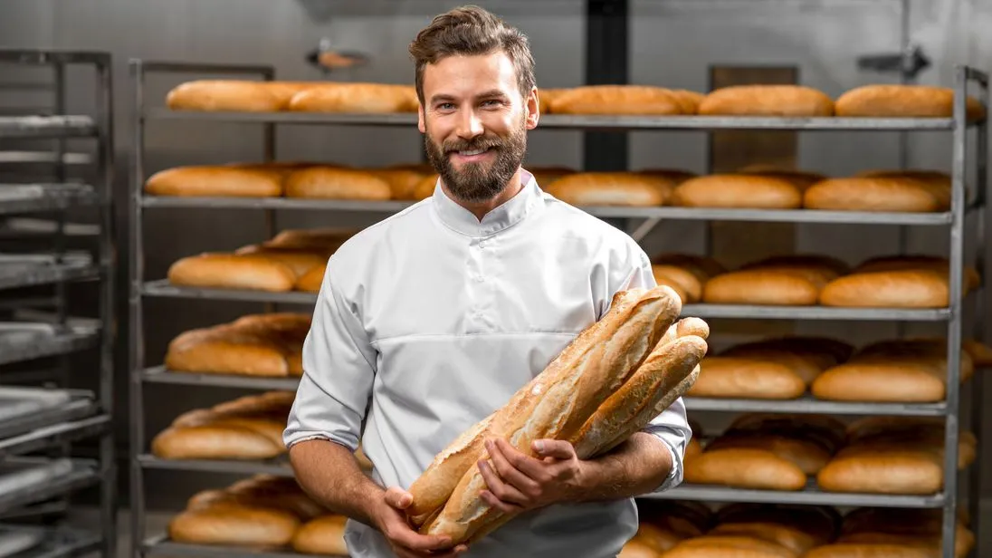 Ein Bäcker steht vor einem Regal mit Broten und hält mehrere Baguette im Arm.