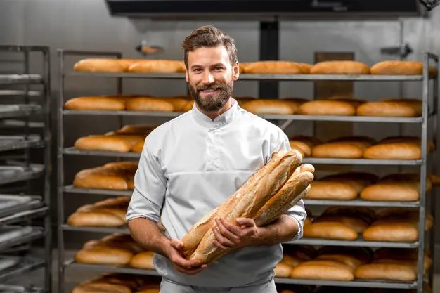 Ein Bäcker steht vor einem Regal mit Broten und hält mehrere Baguette im Arm.