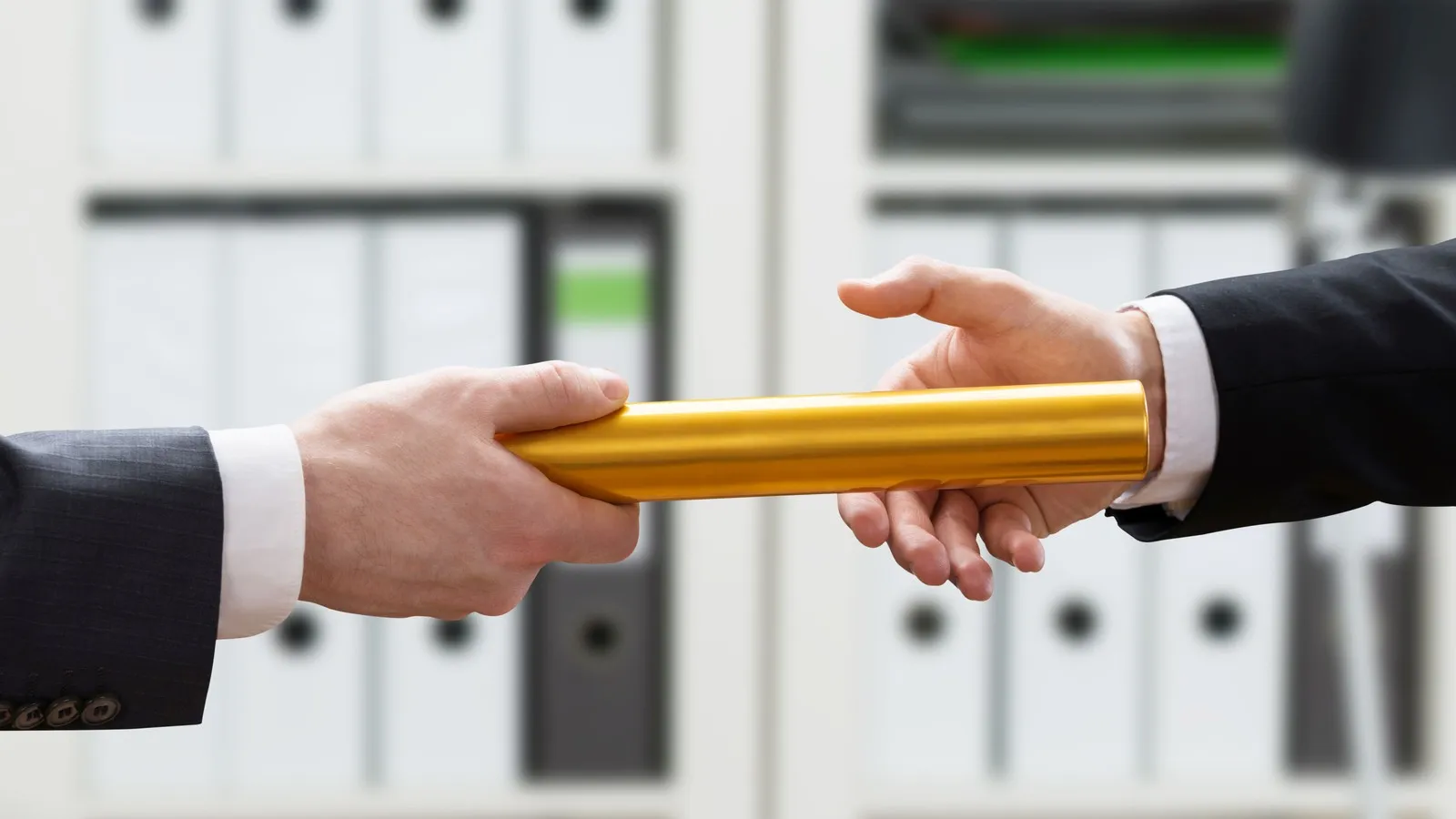 Zwei Männer im Handwerk stehen vor einem Regal, das mit Aktenordnern gefüllt ist. Sie übergeben von Hand zu Hand einen Staffelstab.