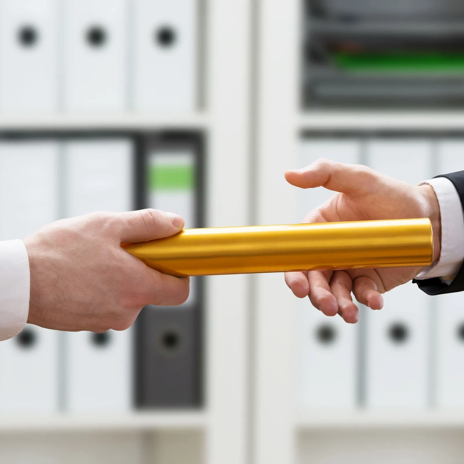 Zwei Männer im Handwerk stehen vor einem Regal, das mit Aktenordnern gefüllt ist. Sie übergeben von Hand zu Hand einen Staffelstab.