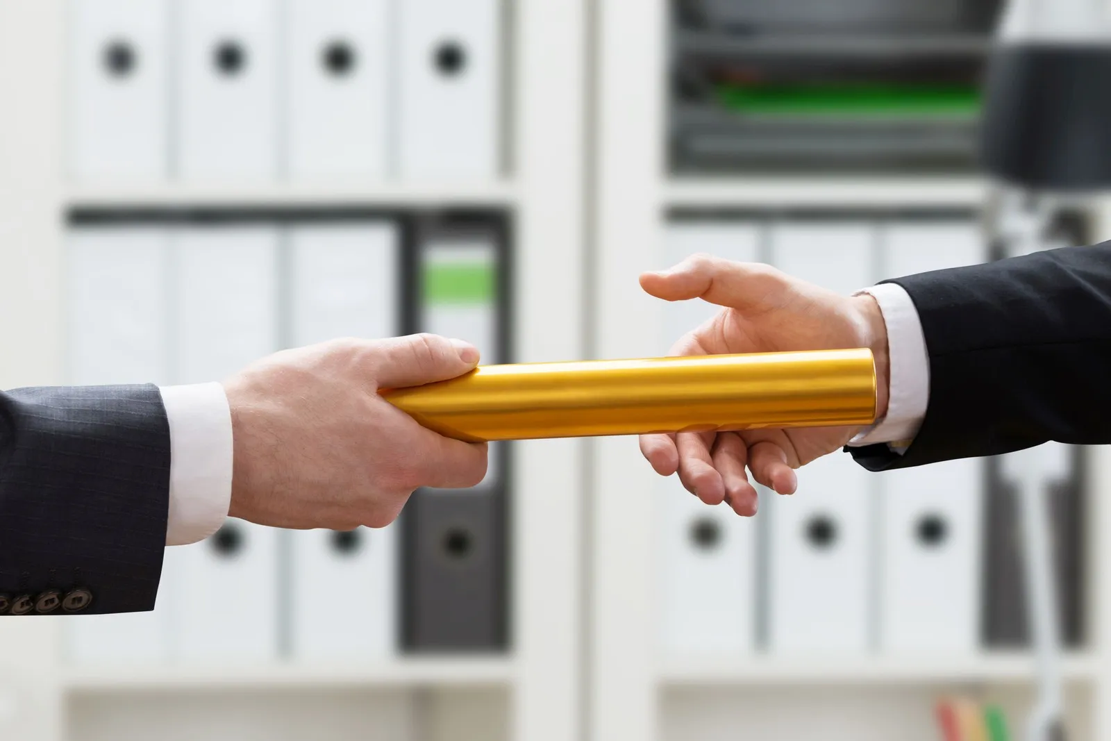 Zwei Männer im Handwerk stehen vor einem Regal, das mit Aktenordnern gefüllt ist. Sie übergeben von Hand zu Hand einen Staffelstab.