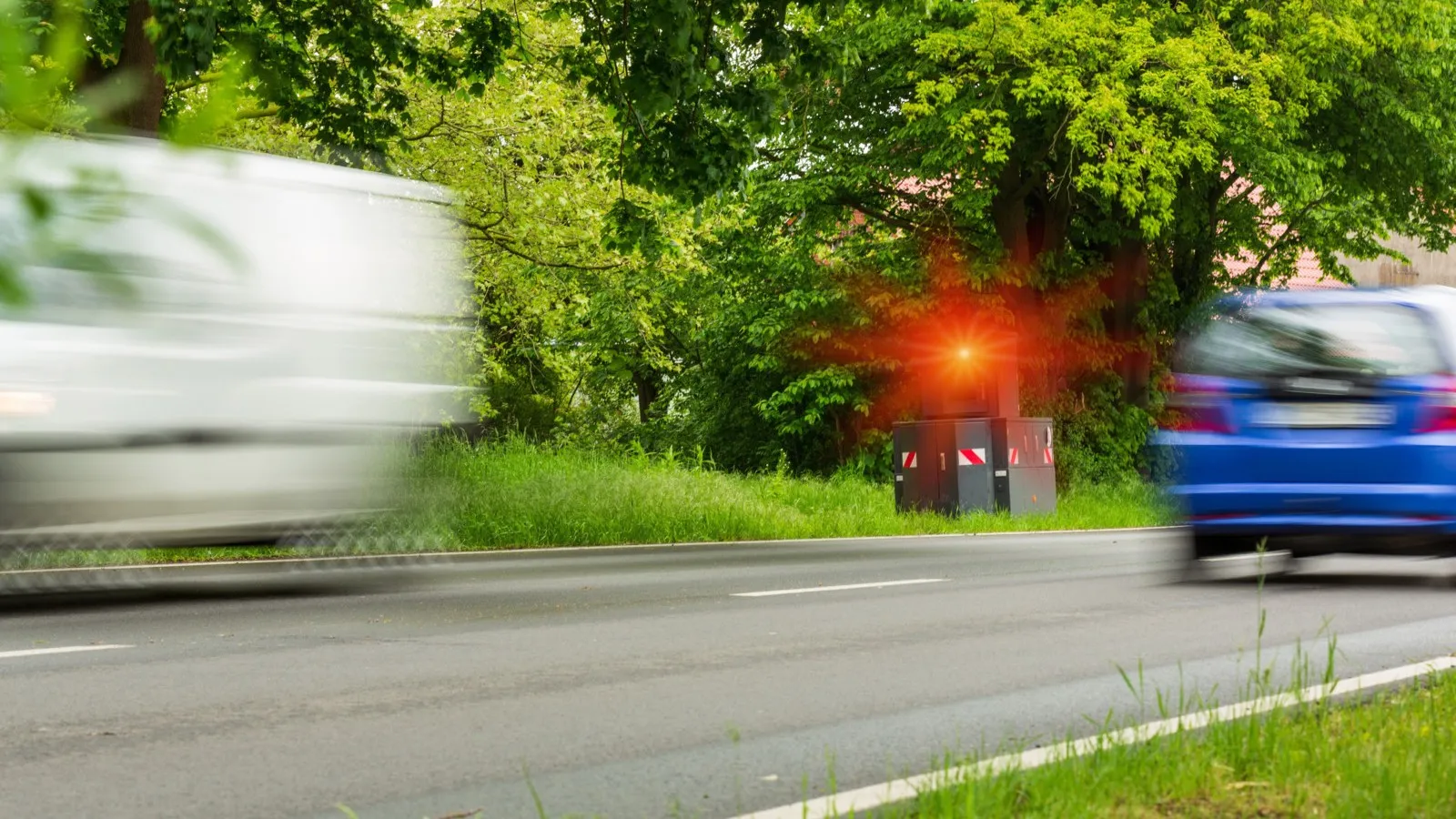 Auf einer befahrenen Straße steht am Rand ein Blitzer.. Es blitzt ein rotes Licht auf, weil die Auto dort zu schnell unterwegs sind.