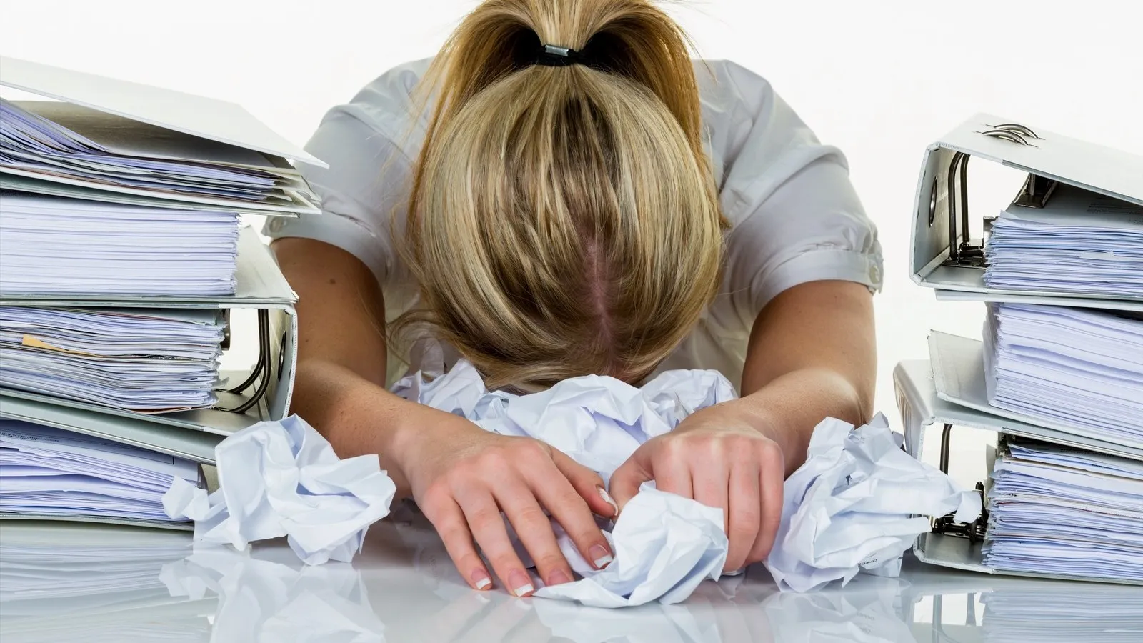 Junge Frau im Büro umgeben von Ordnern ist mit der Arbeit überfordert, sie legt ihren Kopf auf der Tischplatte ab. Burnout bei Arbeit oder Studium.