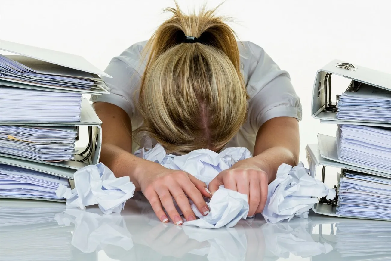 Junge Frau im Büro umgeben von Ordnern ist mit der Arbeit überfordert, sie legt ihren Kopf auf der Tischplatte ab. Burnout bei Arbeit oder Studium.