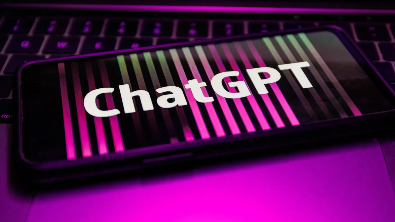 Ein Handy liegt auf einer Laptoptastatur. Fokus liegt auf dem Handy, welches auf dem Bildschirm einen gestreiften schwarz lilanen Hintergrund und der Aufschrift "ChatGPT" zeigt.