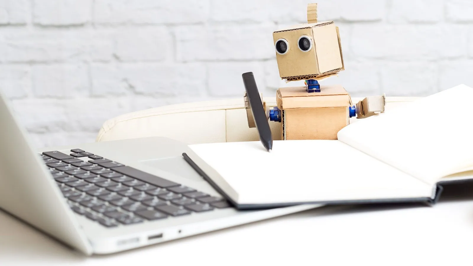 Ein kleiner holzfarbener Roboter hält einen Stift in der Hand und macht sich Notizen in einem Notizheft, während er auf einen Laptop guckt.