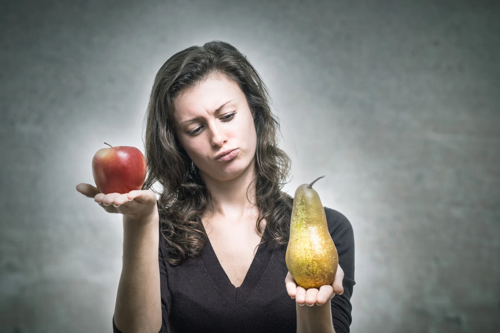Eine Frau mit braunen Locken steht vor einem grauen Hintergrund und hält eine Birne in der einen und einen Apfel in der anderen Hand und macht dabei eine abwägende Geste.