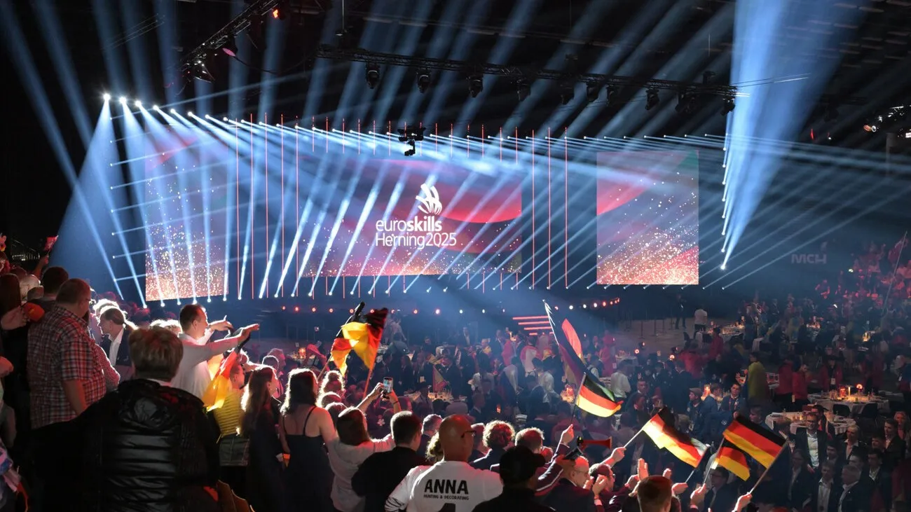 Die Fans der Teilnehmer und Teilnehmerinnen wedeln mit Deutschland-Flaggen. Einige der Personen stehen. Im Hintergrund ist die Bühne zu sehen.