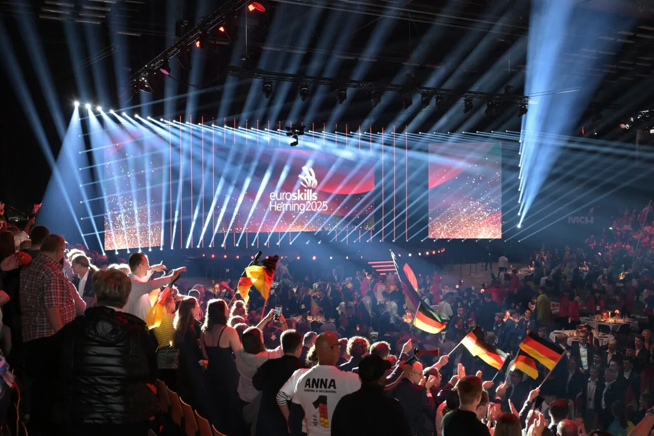 Die Fans der Teilnehmer und Teilnehmerinnen wedeln mit Deutschland-Flaggen. Einige der Personen stehen. Im Hintergrund ist die Bühne zu sehen.