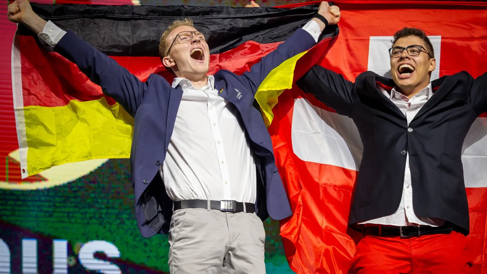 Linus Großhardt (links) trägt ein blaues Jacket und ein weißes Hemd. Der Zimmerer freut sich über seine Goldmedaille bei den Euroskills 2025 und streckt deshalb seine Hände nach oben.