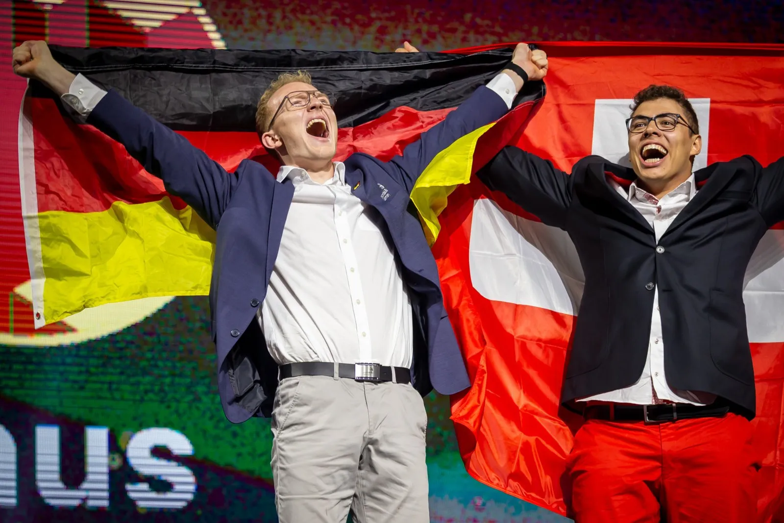Linus Großhardt (links) trägt ein blaues Jacket und ein weißes Hemd. Der Zimmerer freut sich über seine Goldmedaille bei den Euroskills 2025 und streckt deshalb seine Hände nach oben.