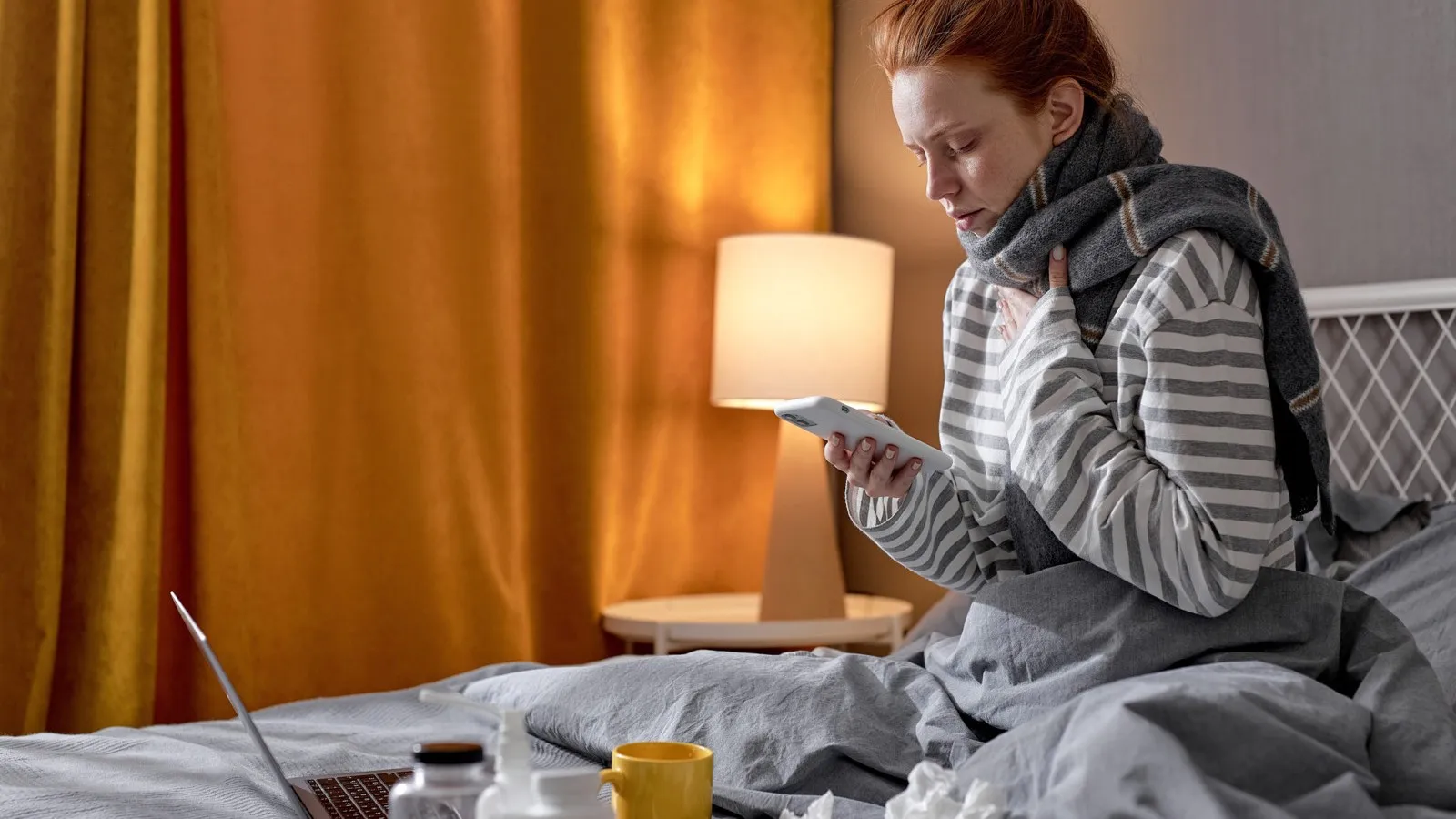 Eine Frau sitzt mit Schal im Bett vor ihrem Laptop und mit dem Smartphone in der Hand. Es soll signalisieren, dass die Frau krank ist.