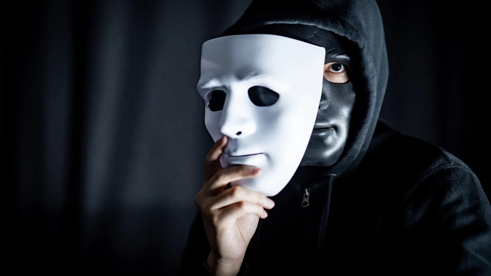Eine schwarz gekleidete Person mit Hoodie und schwarzer Gesichtsmaske hält vor einem schwarzen Hintergrund eine weiße Maske teilweise vor ihr Gesicht.