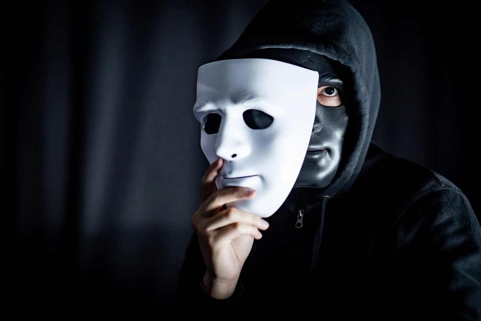 Eine schwarz gekleidete Person mit Hoodie und schwarzer Gesichtsmaske hält vor einem schwarzen Hintergrund eine weiße Maske teilweise vor ihr Gesicht.