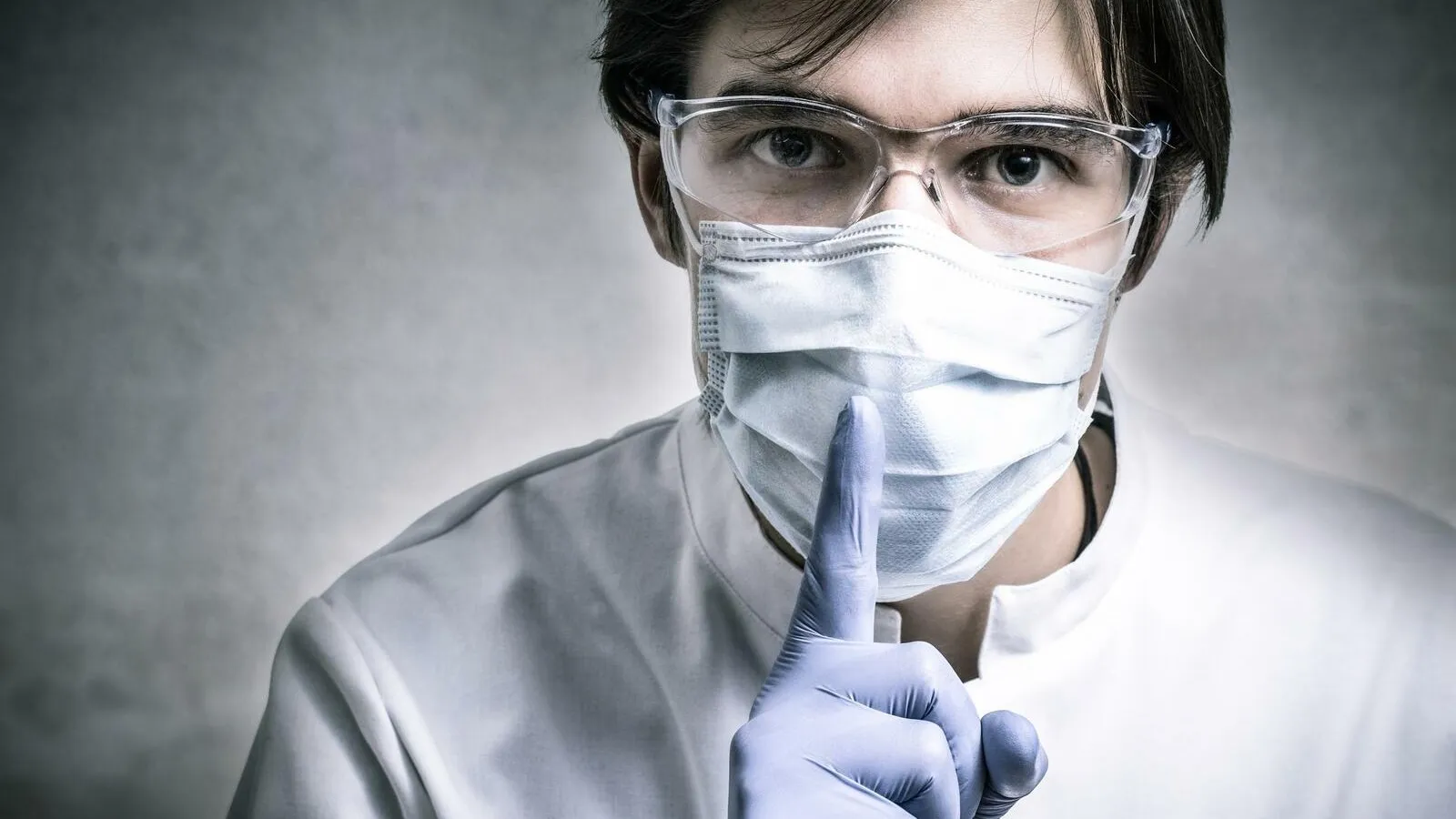 Ein Mann in einer ärztlichen Berufsbekleidung mit Brille und Mundmaske hält sich einen Finger vor den Mund und signalisiert damit Geheimhaltung.