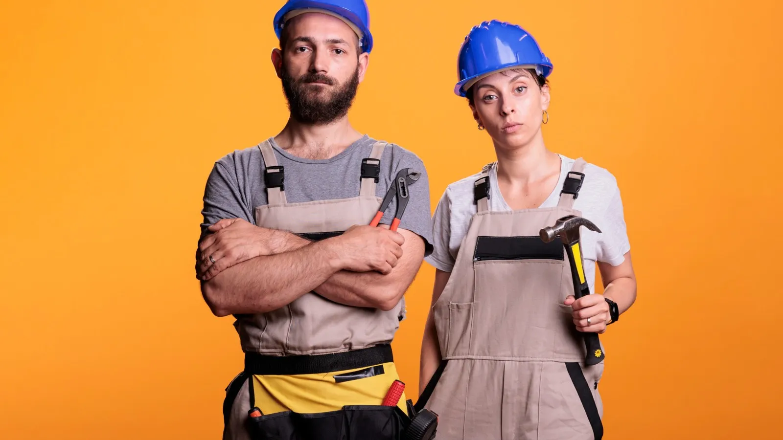 Zwei Handwerker in Latzhose und mit blauem Schutzhelm stehen vor einem orangenen Hintergrund. Sie schauen frustriert.