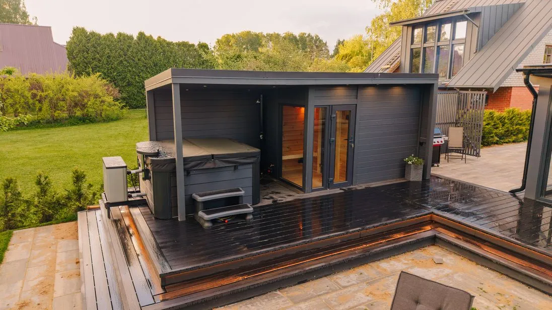 Eine Gartensauna in dunklem Holz steht nahe eines Hauses.