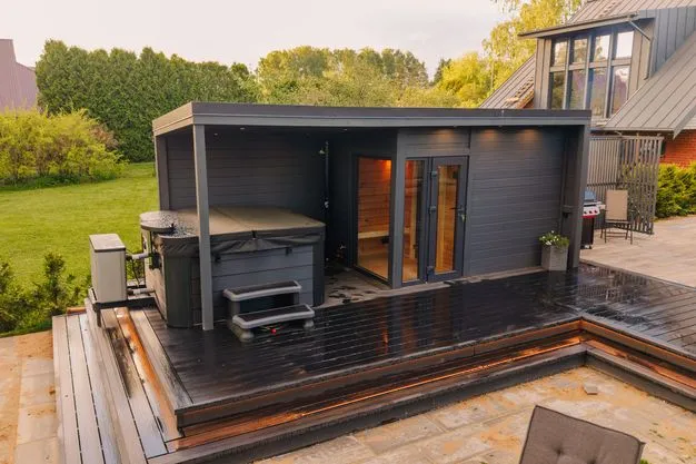 Eine Gartensauna in dunklem Holz steht nahe eines Hauses.