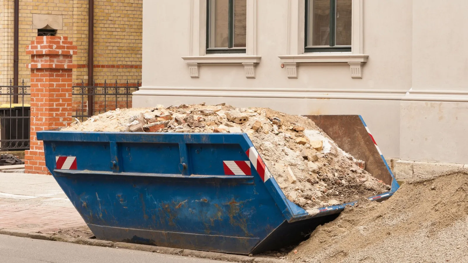 Ein blauer Container gefüllt mit Bauschutt steht vor einem weißen Haus.