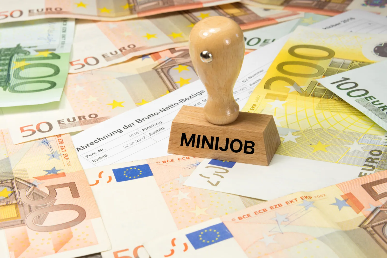 Mehrere Banknoten liegen verteilt, darauf steht ein Holzstempel mit der Aufschrift "Minijob".