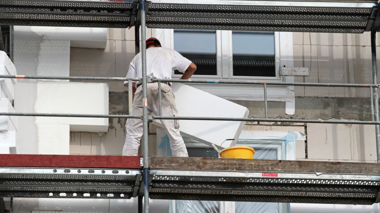 Ein weiß gekleideter Bauhandwerker steht auf einem Gerüst und arbeitet an einem Gebäude.
